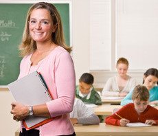 Teacher smiling for the camera while children are studying at their desks.